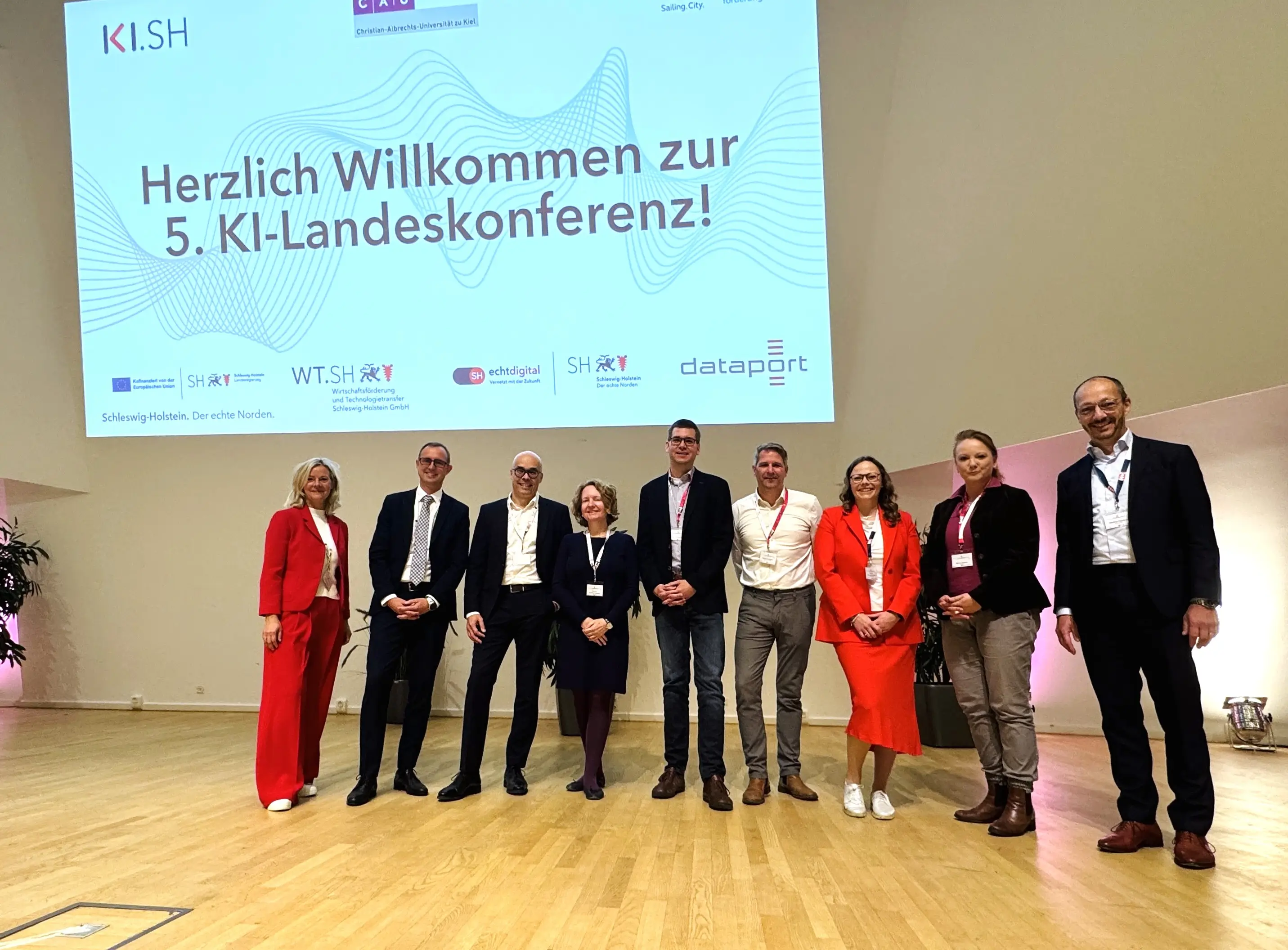 Neun Personen stehen auf einer Bühne vor einem Willkommensgruß zur 5. KI-Landeskonferenz