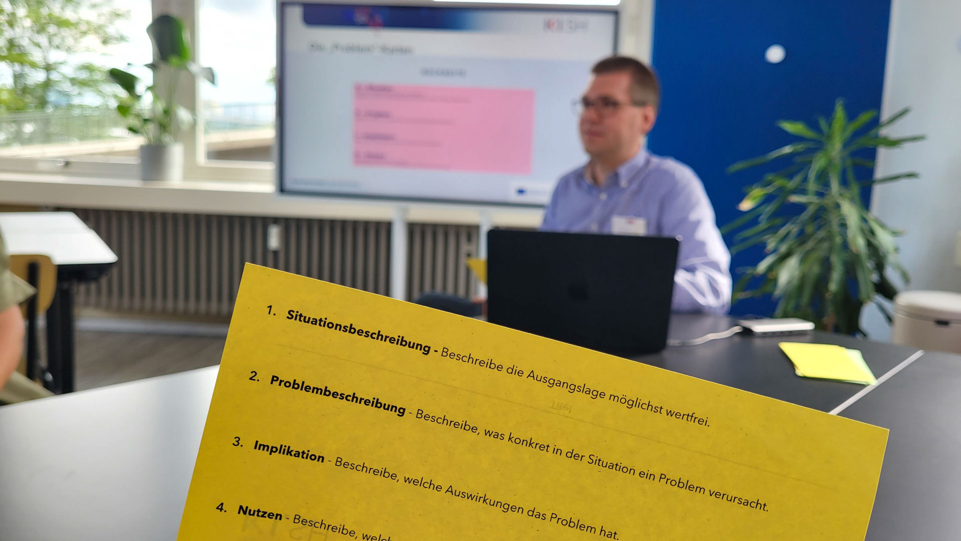 Eine Workshopkarte liegt im Fokus des Bildes. Im Hintergrund sitzt ein Mann vor einem Bildschirm, auf dem die gleiche Karte abgebildet ist.