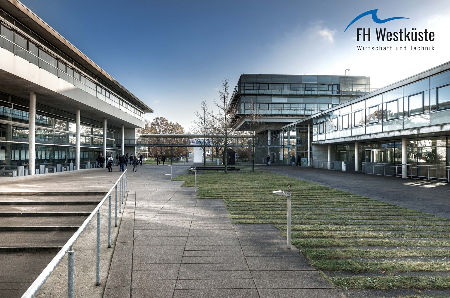 Campus der FH Westküste. Zwei Gebäude mit Glasfassaden und Betonsäulen stehen sich gegenüber. Links führt eine Treppe aus dem Bild. Oben in der Ecke rechts steht das Logo der FH Westküste.