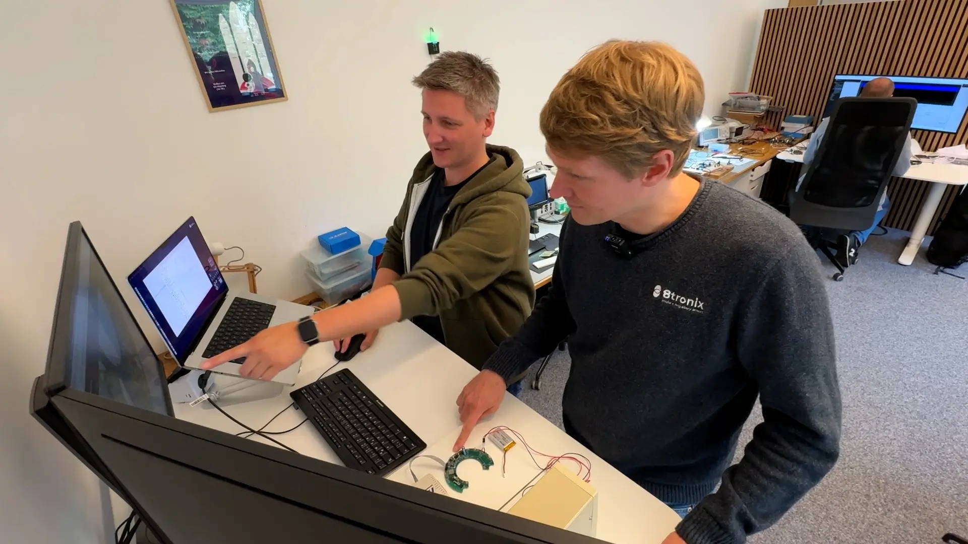 Zwei Personen arbeiten gemeinsam an einem Schreibtisch vor Monitoren und Laptops, zeigen auf den Bildschirm und diskutieren eine elektronische Schaltung, im Hintergrund sitzt eine weitere Person an einem Arbeitsplatz..